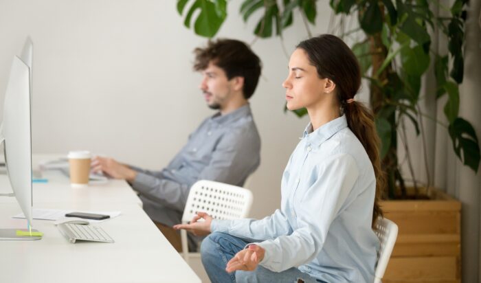 Young calm woman taking break in office for meditation, mindful peaceful millennial girl employee or student practicing yoga for relaxation sitting in lotus pose, stress free relief at work concept